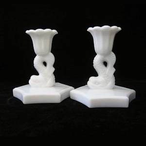 May include: Pair of white candle holders. Each holder features a dolphin-shaped base supporting a fluted, flower-like cup. The candle holders are made of a solid white material and sit on a square base. The background is black.