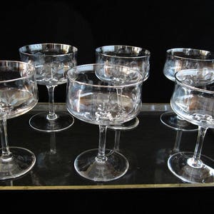 May include: Six clear champagne coupe glasses with silver rims are arranged on a black tray with gold trim. The glasses have a classic design with a wide bowl and a slender stem. The background is a solid black.
