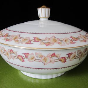 May include: A white porcelain serving bowl with a lid, featuring a floral design in pink and gold. The bowl has a gold rim and handles, and a decorative finial on the lid. The bowl is likely for serving food.