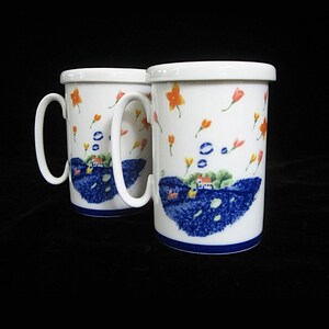 May include: Two white ceramic mugs with a whimsical design. Each mug features a blue and white landscape scene with a house, blue water, and orange and yellow flowers. The mugs have white handles and a blue trim.