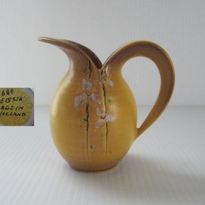 May include: A small, yellow ceramic pitcher with a brown handle and floral design. The pitcher has a label on the bottom that reads "689 Deysja Made in Holland".