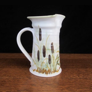May include: White ceramic pitcher with a green rim and a brown and green floral design. The pitcher has a handle and a spout.