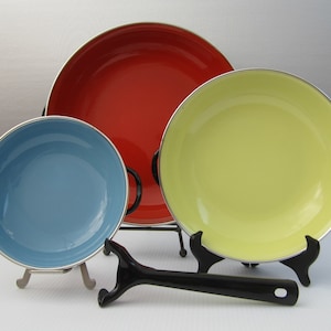 May include: Three enamelware bowls in different sizes, red, yellow, and blue, on a white surface. The bowls are on metal stands and there is a black utensil in the foreground.