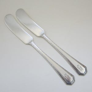 May include: Two silver butter knives with ornate handles. The handles have a repeating pattern and a monogrammed initial.