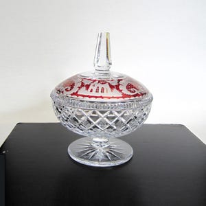 May include: A clear crystal candy dish with a lid. The lid has a red interior with a white etched design. The dish has a diamond pattern and sits on a round base. The finial on the lid is clear and elongated.