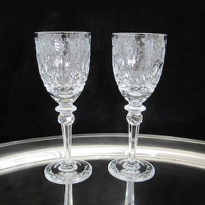 May include: Two clear crystal wine glasses with etched floral and figure designs. The glasses have a diamond-cut pattern on the bowl, a decorative stem, and a round base. The glasses are displayed on a silver tray against a black background.