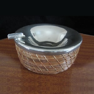 May include: A silver ashtray with a woven wicker base. The ashtray has a round, shallow bowl and a small, rectangular slot for cigarettes.