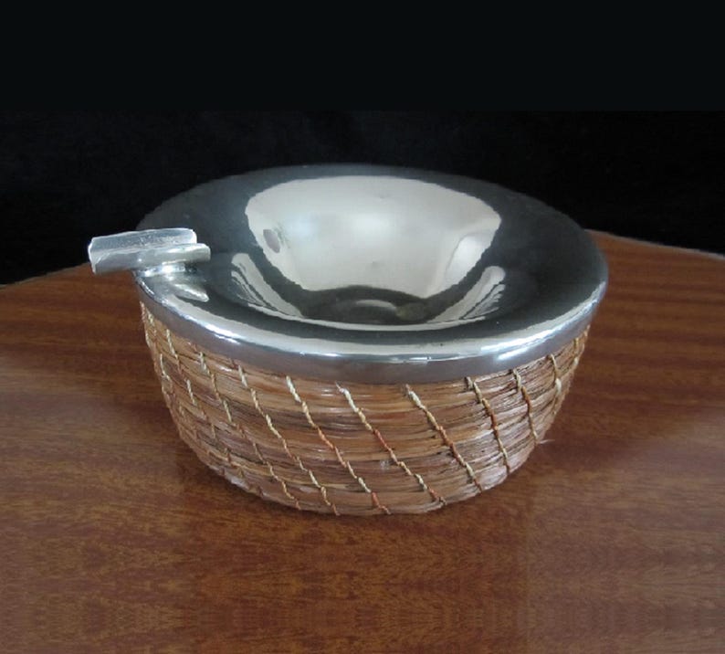 May include: A silver ashtray with a woven wicker base. The ashtray has a round, shallow bowl and a small, rectangular slot for cigarettes.