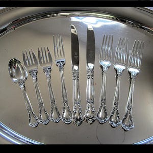 May include: An assortment of silver-colored silverware, including forks, knives, and a spoon, arranged on a silver serving tray. The silverware features ornate, decorative handles. The arrangement suggests a formal dining setting.