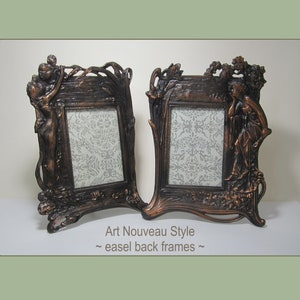 May include: A pair of antique-style, bronze-colored picture frames with an Art Nouveau design. Each frame features a sculpted female figure and intricate floral details. The frames have easel backs for tabletop display.