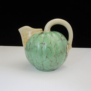 May include: A green ceramic pitcher with a textured surface and a cream-colored handle. The pitcher is shaped like a gourd.