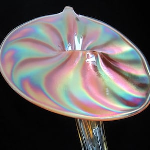 May include: A iridescent glass vase with a swirling, flower-shaped opening. The vase is a pale pink color with a pearlescent finish.
