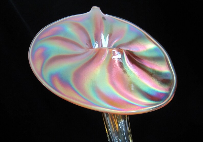 May include: A iridescent glass vase with a swirling, flower-shaped opening. The vase is a pale pink color with a pearlescent finish.