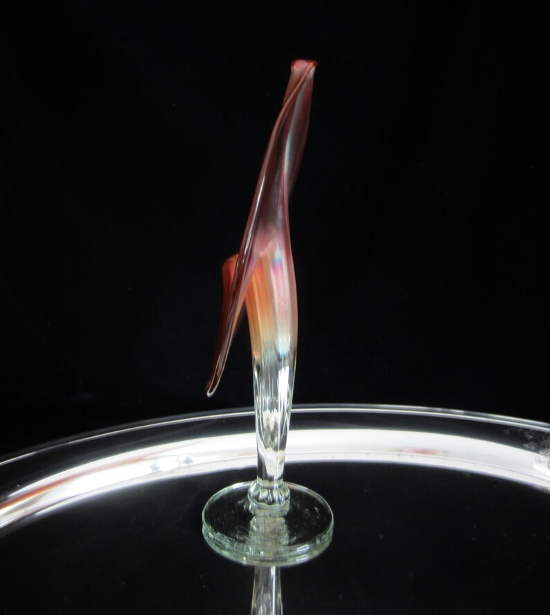 May include: A glass sculpture of a flower with a long, thin stem and a single, large, red and orange petal. The sculpture is on a clear glass base.