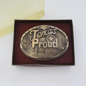 May include: A gold-toned belt buckle with an oval shape and a raised design. The buckle features a cowboy riding a horse, an oil derrick, a building, and the text "Texas Proud".