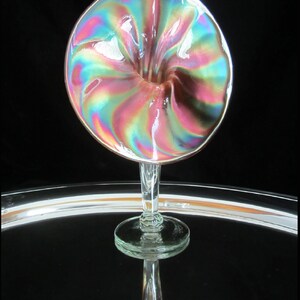 May include: A iridescent glass vase in the shape of a flower with a clear glass stem and base. The vase has a pink, green, and blue iridescent finish.