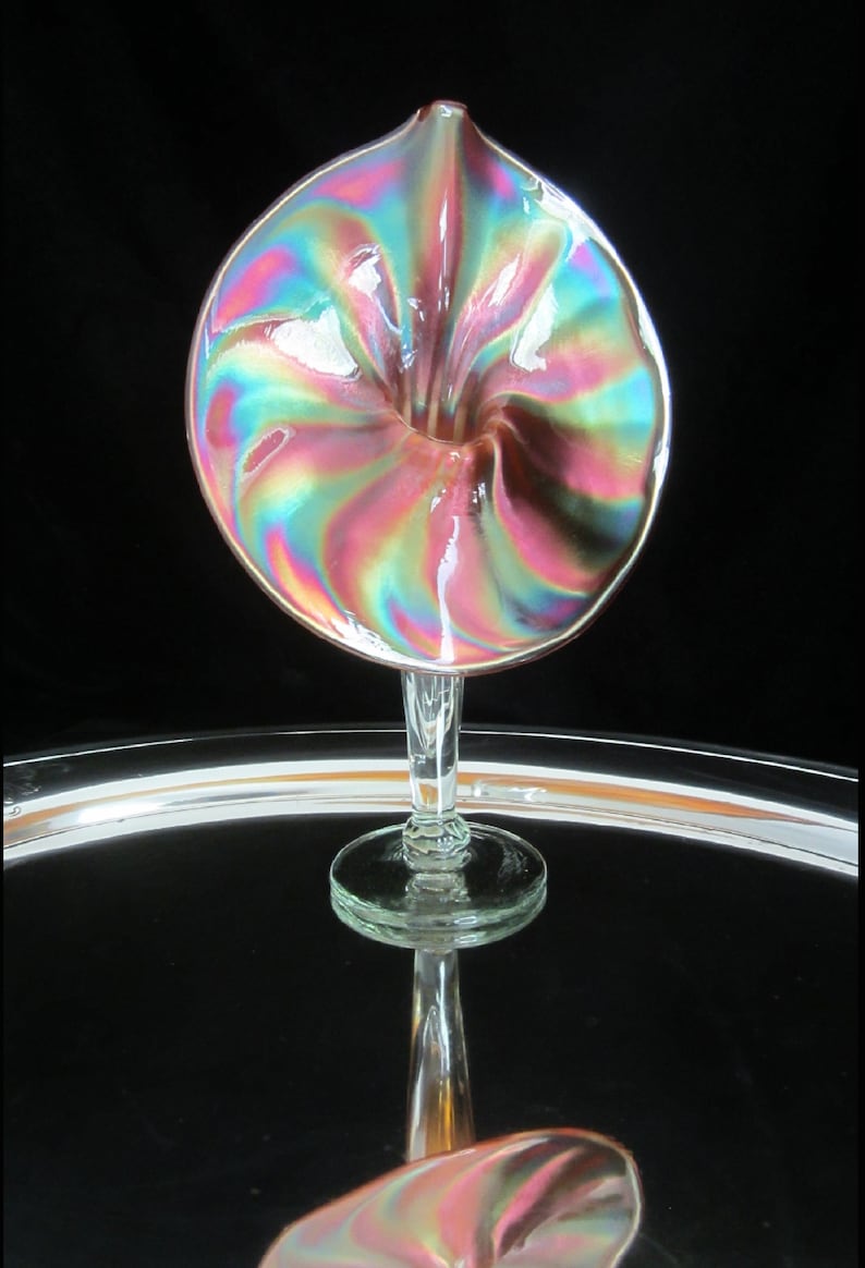 May include: A iridescent glass vase in the shape of a flower with a clear glass stem and base. The vase has a pink, green, and blue iridescent finish.