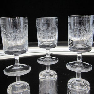 May include: Three clear glass shot glasses with etched floral designs. The glasses have a short stem and a wide base.