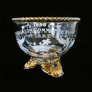 May include: A clear glass bowl with a gold rim and base. The bowl has decorative white lettering and designs, including the text "1898 ASCALON COMMANDERY No. 59, K.T." The base is ornate with gold detailing.