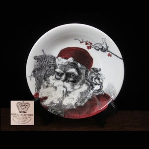 May include: A white ceramic plate with a red and black illustration of Santa Claus. The plate has a red rim and a black and white illustration of a bird on a branch with red berries.