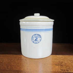 May include: A white ceramic crock with a lid, featuring a blue stripe and a circular emblem that reads "Garden City Potter" with the number "2". The antique container is likely used for food storage.