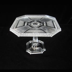 May include: A clear glass cake stand with a square base and a geometric design on the top plate.