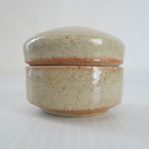 May include: A small, two-piece ceramic container with a lid. The container is a light beige color with a subtle speckled texture. The lid has a slightly raised edge.