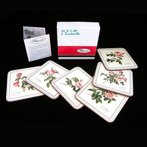 May include: A set of square coasters with a floral rose design. The coasters feature a cream background with pink roses and green leaves. The set includes a white box with red trim and a small pamphlet.