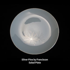 May include: A light blue salad plate with a gold rim and a stylized pine cone design in the center.
