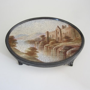 May include: An oval ceramic trinket dish with a silver rim and three feet. The dish features a painted scene of a castle with a waterfall in the background.