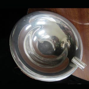 May include: A silver ashtray with a rounded, bowl-shaped design. The ashtray has a smooth, reflective surface and a small, curved lip on one side.