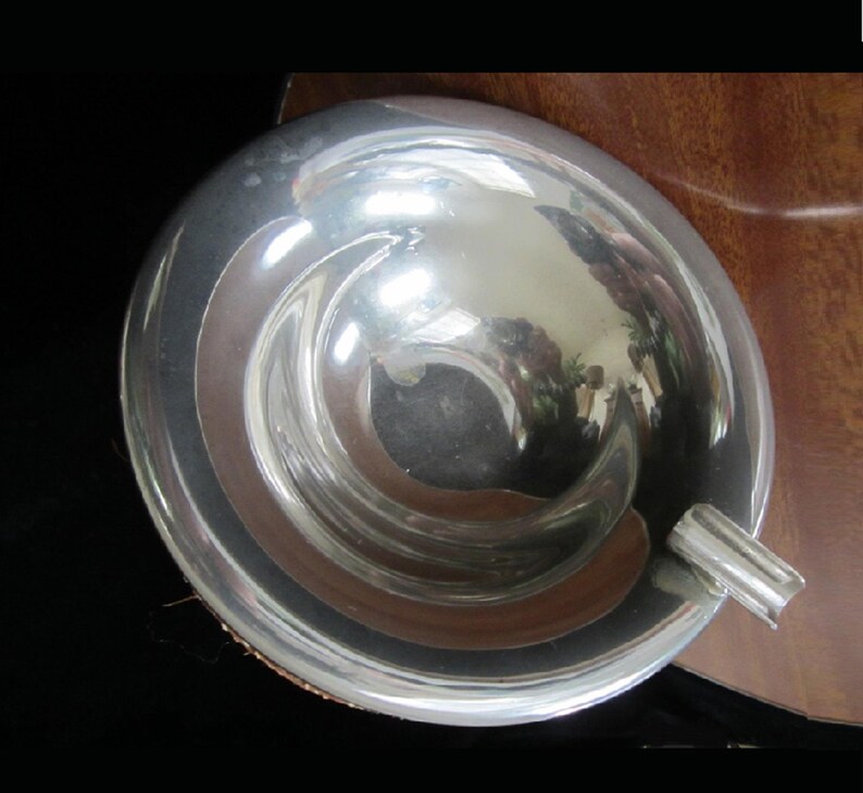 May include: A silver ashtray with a rounded, bowl-shaped design. The ashtray has a smooth, reflective surface and a small, curved lip on one side.