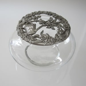 May include: A clear glass jar with a silver lid featuring a floral design with a watering can. The lid is ornate and detailed.