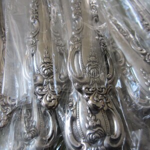May include: A close-up of a set of silver flatware with ornate handles. The handles are decorated with a floral design and are wrapped in clear plastic.
