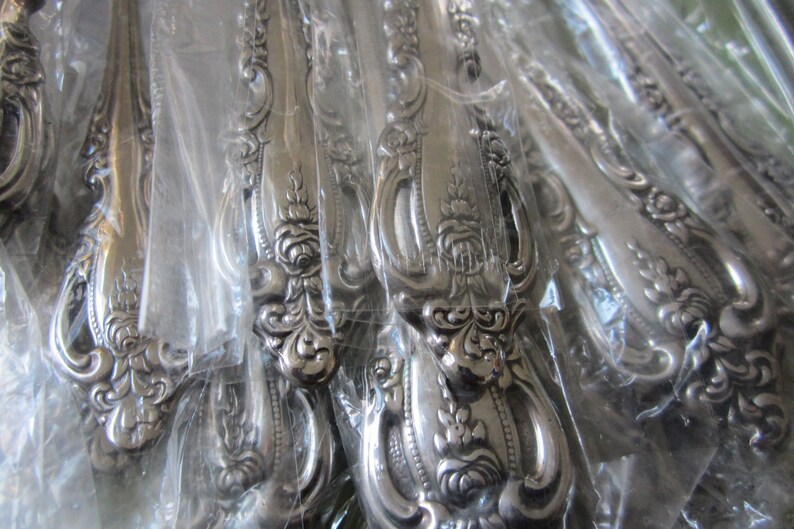May include: A close-up of a set of silver flatware with ornate handles. The handles are decorated with a floral design and are wrapped in clear plastic.