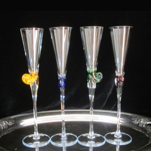May include: Four clear glass champagne flutes with colorful glass accents on the stems. The accents are yellow, blue, green, and burgundy.
