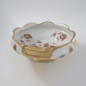 May include: A small, white porcelain bowl with a scalloped rim and gold trim. The bowl is decorated with a floral pattern in shades of purple, brown, and green.