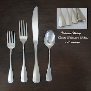 May include: A set of five silver-plated flatware pieces, including a knife, fork, teaspoon, and two dinner forks. The flatware is part of the Oneida Distinction Deluxe 1975 pattern, known as Colonial Artistry.
