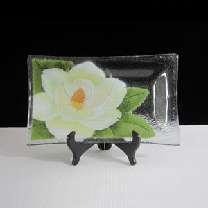 May include: A clear glass rectangular dish with a white magnolia flower and green leaves painted on the bottom. The dish is on a black stand.