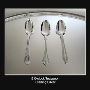May include: Three sterling silver teaspoons with ornate handle designs are arranged on a reflective silver tray. The spoons are labeled "5 O'clock Teaspoon" and are highly polished, reflecting the light. The spoons are elegant and suitable for serving tea.