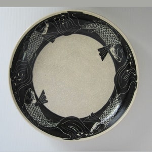May include: A white ceramic plate with a black border and a design of four fish swimming in a circle.