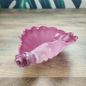 May include: A decorative glass bowl shaped like a conch shell, featuring a gradient of translucent light pink to a deeper, darker purple at the spiral tip. The bowl has a ruffled, scalloped edge and a smooth, glossy finish throughout the piece.