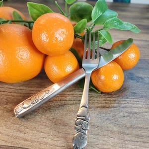 May include: A still life featuring bright orange tangerines and green leaves arranged on a wooden surface. A silver fork and knife with decorative handles are placed among the fruit. The silverware has a floral and bunny design.