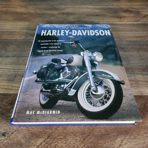 May include: A hardcover book titled The Ultimate HARLEY-DAVIDSON by Mac McDiarmid. The cover features a photograph of a vintage-style motorcycle with white-wall tires, chrome accents, and a black leather saddlebag parked on a gravel road.