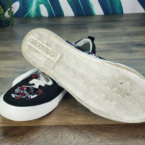 May include: A pair of black slip-on sneakers featuring a white rubber sole with a herringbone tread pattern and the word GROUND UP embossed on the bottom. The upper fabric displays a colorful repeating pattern of cartoon character faces.