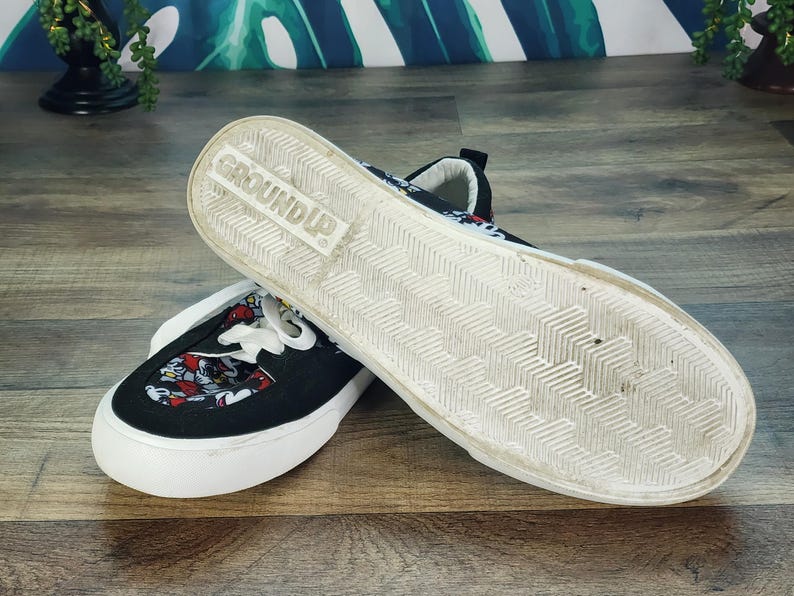May include: A pair of black slip-on sneakers featuring a white rubber sole with a herringbone tread pattern and the word GROUND UP embossed on the bottom. The upper fabric displays a colorful repeating pattern of cartoon character faces.