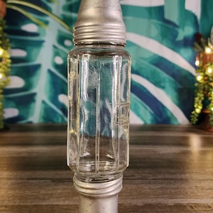 May include: A clear glass cocktail shaker with a ribbed design and a silver metal top and base. The shaker has a conical top and a cylindrical body. The metal components show signs of wear, giving it a vintage appearance.
