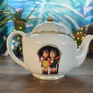 May include: White ceramic teapot with a lid and a gold-colored handle and trim. The teapot features a painted image of two children under an umbrella. The background is a blurred image of plants.