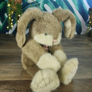 Könnte beinhalten: Ein Plüsch-Hasen-Spielzeug mit weichem, hellbraunem Fell und cremefarbenen Akzenten an Ohren, Pfoten und Bauch. Der Hase hat eine rosa Nase und sitzt aufrecht. Das Spielzeug steht auf einer Holzoberfläche.