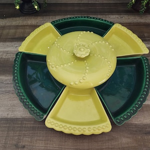 May include: A circular ceramic serving platter featuring a central yellow lidded bowl surrounded by alternating dark green and light yellow wedge-shaped dishes. Each dish has a beaded edge, and the lid features a raised spiral dot pattern.